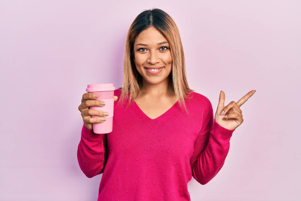Bella donna ispanica bere una tazza di caffè da asporto sorridente felice indicando con mano e dito di lato  - Foto, immagini