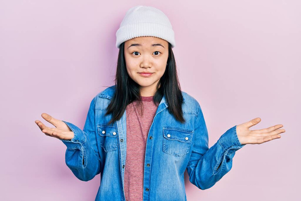 Fiatal kínai lány visel aranyos gyapjú sapka tanácstalan és zavaros kifejezés karok és kezek emelt. kétség.  - Fotó, kép