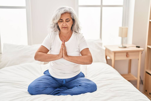 Middle age woman doing yoga exercise sitting on bed at bedroom - 写真・画像