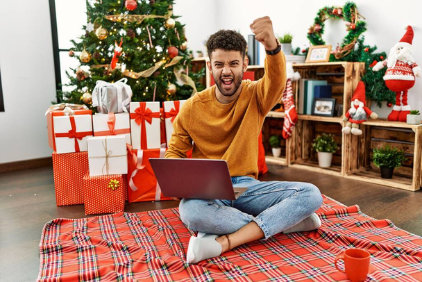 Arab fiatalember használja laptop ül karácsonyfa dühös és őrült emelő ököl frusztrált és dühös, miközben kiabál a harag. düh és agresszív koncepció.  - Fotó, kép