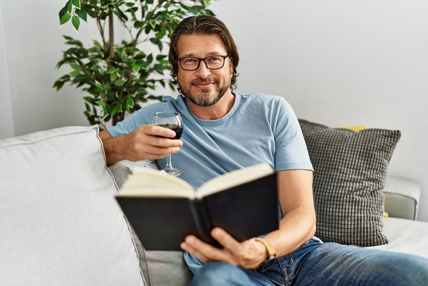 Middle age caucasian man reading book drinking wine at home - Photo, Image