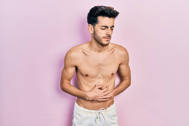 Νεαρός Ισπανός που φοράει μαγιό χωρίς πουκάμισο με το χέρι στο στομάχι επειδή η δυσπεψία, η επώδυνη ασθένεια δεν αισθάνεται καλά. έννοια του πόνου.  - Φωτογραφία, εικόνα