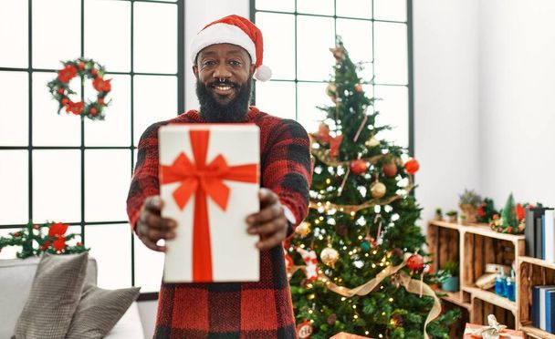 Молодой африканский американец с подарком стоит у рождественской елки дома - Фото, изображение