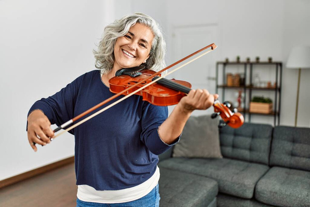 Middle age grey-haired artist woman playing violin standing at home. - Fotografie, Obrázek
