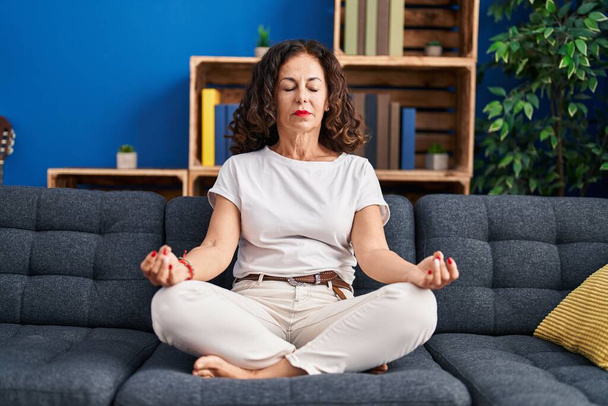 Střední věk hispánská žena dělá jógu pózování sedí na pohovce doma - Fotografie, Obrázek