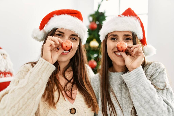 Two women smiling confident with christmas ball on nose at home - 写真・画像