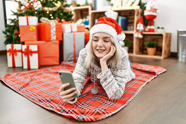 Νεαρή λευκή κοπέλα που φοράει χριστουγεννιάτικο καπέλο και φτιάχνει σέλφι δίπλα στο smartphone που είναι ξαπλωμένο στο πάτωμα του σπιτιού. - Φωτογραφία, εικόνα