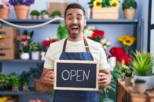 Hombre hispano con barba trabajando en floristería sosteniendo letrero abierto sonriendo y riendo en voz alta porque gracioso chiste loco.  - Foto, imagen