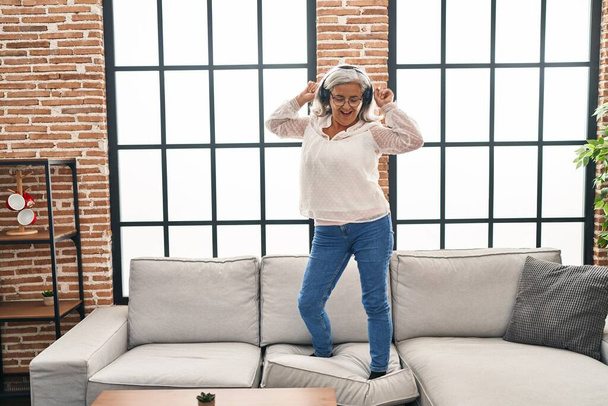 Μέση ηλικία γυναίκα ακούγοντας μουσική και χορεύοντας στέκεται στον καναπέ στο σπίτι - Φωτογραφία, εικόνα