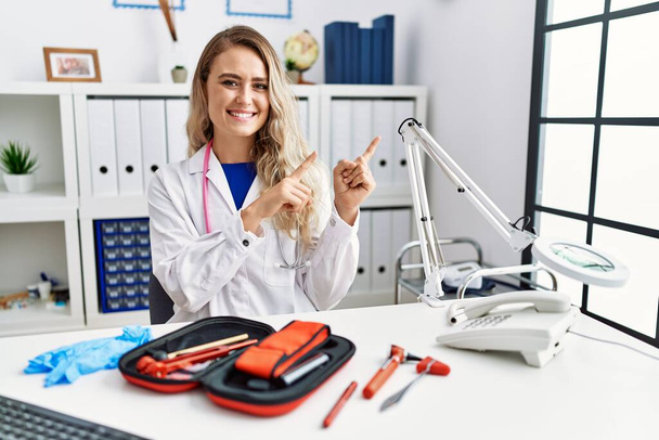 Jonge mooie arts vrouw met reflex hamer en medische instrumenten glimlachen en kijken naar de camera wijzen met twee handen en vingers naar de zijkant.  - Foto, afbeelding