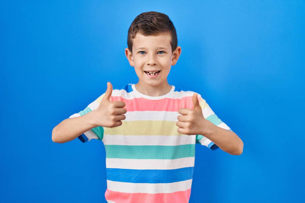 Young caucasian kid standing over blue background success sign doing positive gesture with hand, thumbs up smiling and happy. cheerful expression and winner gesture.  - Zdjęcie, obraz