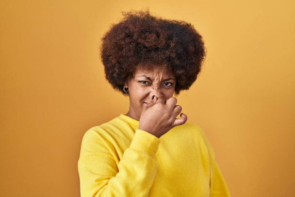Young african american woman standing over yellow background smelling something stinky and disgusting, intolerable smell, holding breath with fingers on nose. bad smell  - Photo, Image