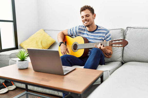 Joven hombre hispano sonriendo confiado tocando concierto de guitarra en línea en casa - Foto, Imagen