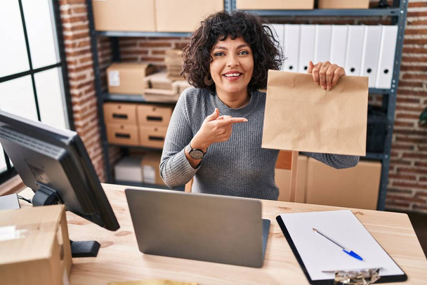 Hispanic doctor woman with curly hair working at small business ecommerce holding paper bag smiling happy pointing with hand and finger  - 写真・画像