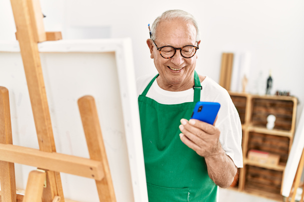 Homem artista de cabelos grisalhos sênior sorrindo feliz e usando pintura smartphone no estúdio de arte. - Foto, Imagem