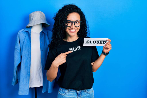Young hispanic woman with curly hair wearing staff t shirt holding closed banner surprised with hand on head for mistake, remember error. forgot, bad memory concept.  - Zdjęcie, obraz