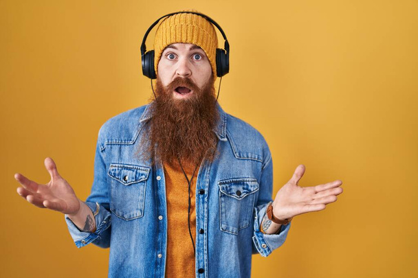 Caucasian man with long beard listening to music using headphones clueless and confused with open arms, no idea concept.  - Fotó, kép
