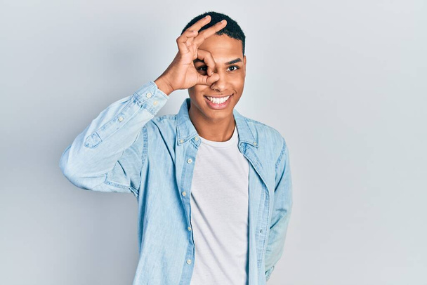 Young african american guy wearing casual clothes doing ok gesture with hand smiling, eye looking through fingers with happy face.  - Фото, изображение