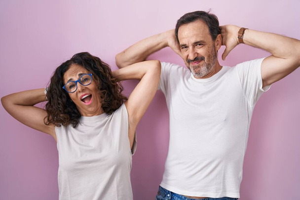Middle age hispanic couple together over pink background relaxing and stretching, arms and hands behind head and neck smiling happy  - Photo, image