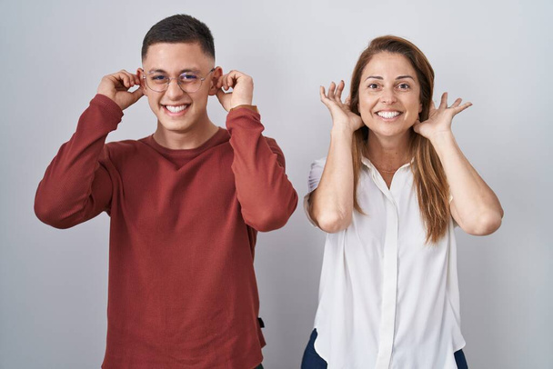 Mother and son standing together over isolated background smiling pulling ears with fingers, funny gesture. audition problem  - Photo, Image