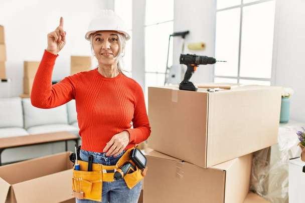 Middle age grey-haired woman wearing hardhat standing at new home pointing finger up with successful idea. exited and happy. number one.  - Foto, imagen