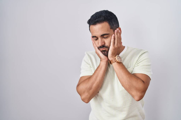 Homme hispanique avec barbe debout sur fond isolé mains fatiguées couvrant le visage, la dépression et la tristesse, contrarié et irrité par le problème  - Photo, image