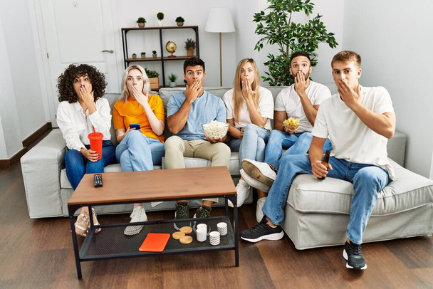 Group of people eating snack sitting on the sofa at home covering mouth with hand, shocked and afraid for mistake. surprised expression  - Foto, immagini