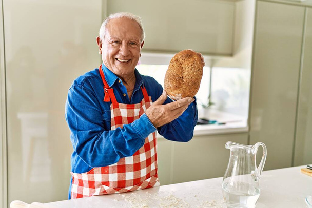 Ηλικιωμένος άνδρας χαμογελά σίγουρος κρατώντας ψωμί ολικής άλεσης στην κουζίνα - Φωτογραφία, εικόνα