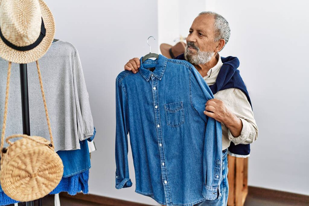 Hombre mayor de pelo gris sonriendo confiado sosteniendo la camisa en la tienda de ropa - Foto, imagen