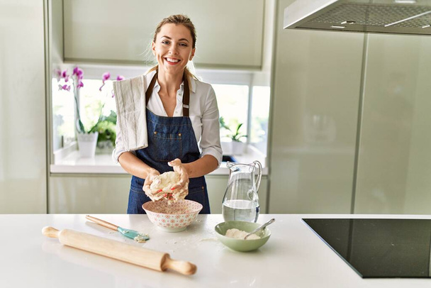Nuori blondi nainen hymyilee luottavainen tehdä pizza taikina kädet keittiössä - Valokuva, kuva
