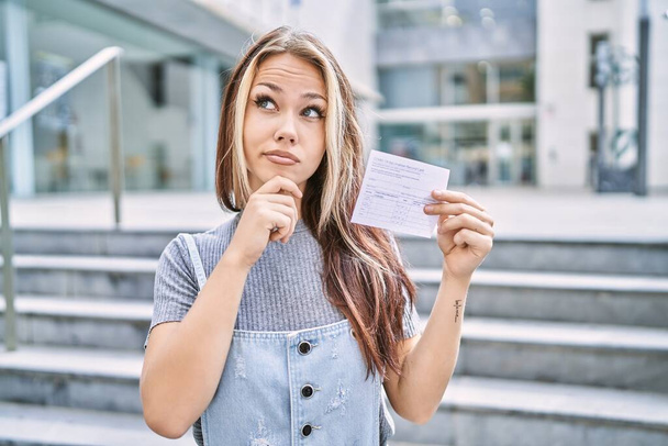 Νεαρή καυκάσια γυναίκα που κρατά κάρτα ρεκόρ covid σοβαρό πρόσωπο σκέφτεται την ερώτηση με το χέρι στο πηγούνι, στοχαστική για σύγχυση ιδέα  - Φωτογραφία, εικόνα