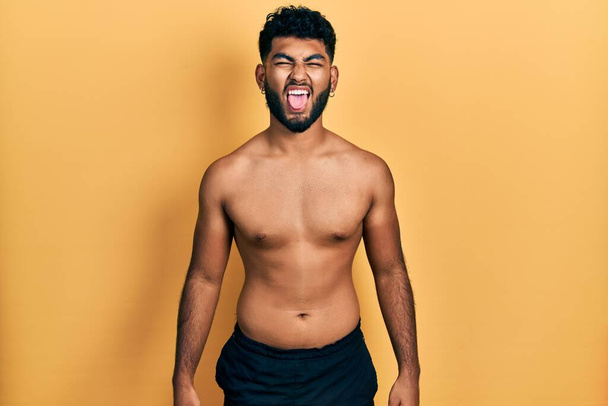 Arab man with beard wearing swimwear shirtless sticking tongue out happy with funny expression. emotion concept.  - 写真・画像