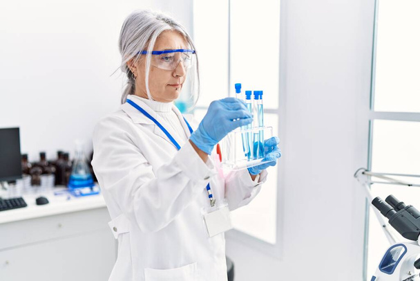 Middle age grey-haired woman wearing scientist uniform using laptop holding test tube at laboratory - Photo, Image