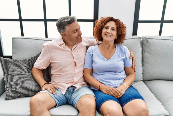 Middelbare leeftijd man en vrouw paar glimlachen zelfverzekerd knuffelen elkaar zitten op de bank thuis - Foto, afbeelding