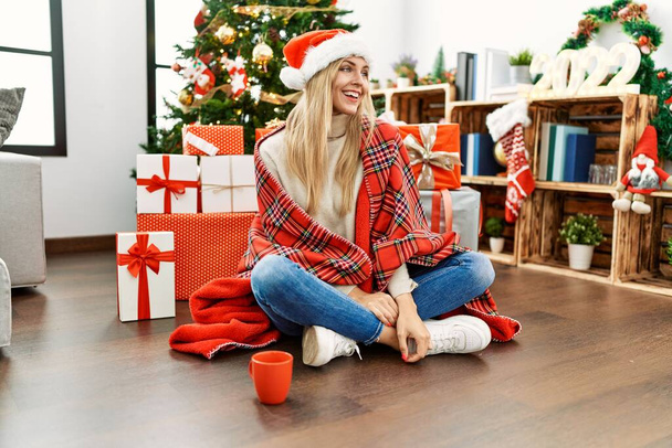 Beautiful blonde woman sitting on the floor by christmas tree looking away to side with smile on face, natural expression. laughing confident.  - Foto, imagen