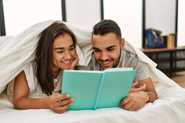 Giovane coppia latina lettura libro copertura con lenzuolo sul letto. - Foto, immagini