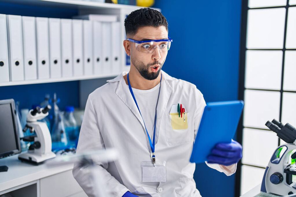 Young handsome man working at scientist laboratory doing online call scared and amazed with open mouth for surprise, disbelief face  - Photo, Image
