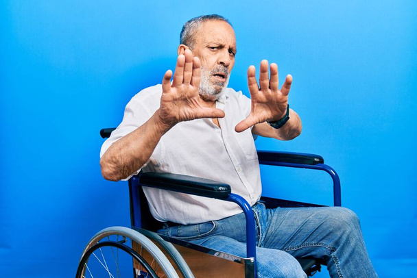 Handsome senior man with beard sitting on wheelchair doing stop gesture with hands palms, angry and frustration expression  - Foto, Imagen