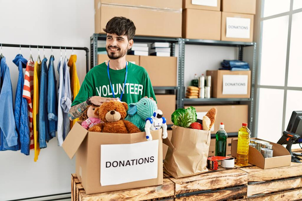Giovane uomo ispanico che indossa uniforme volontaria con scatola di donazioni con giocattoli al centro di beneficenza. - Foto, immagini