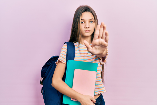 Młoda brunetka dziewczyna gospodarstwa studencki plecak i książki z otwartą ręką robi znak stop z poważnym i pewnym siebie wyrazem, gest obrony  - Zdjęcie, obraz