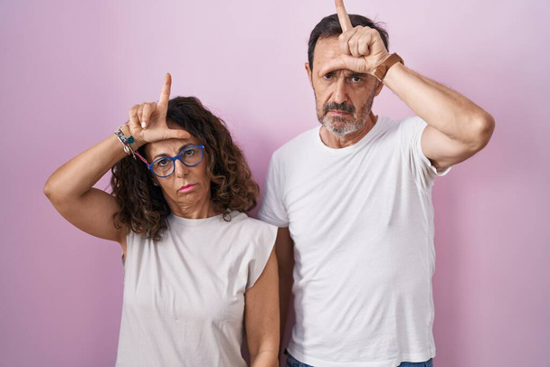 Middle age hispanic couple together over pink background making fun of people with fingers on forehead doing loser gesture mocking and insulting.  - Photo, Image
