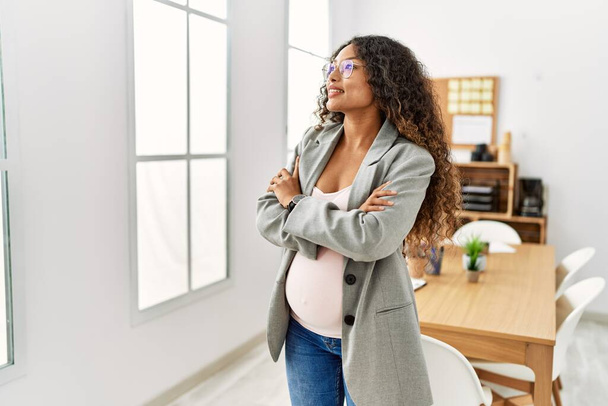 Mladý latinsky žena těhotná usměvavý sebevědomý stojící s rukama zkřížené gesto v kanceláři - Fotografie, Obrázek