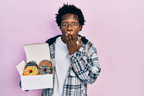 Young african american man holding tasty colorful doughnuts box covering mouth with hand, shocked and afraid for mistake. surprised expression  - Photo, Image