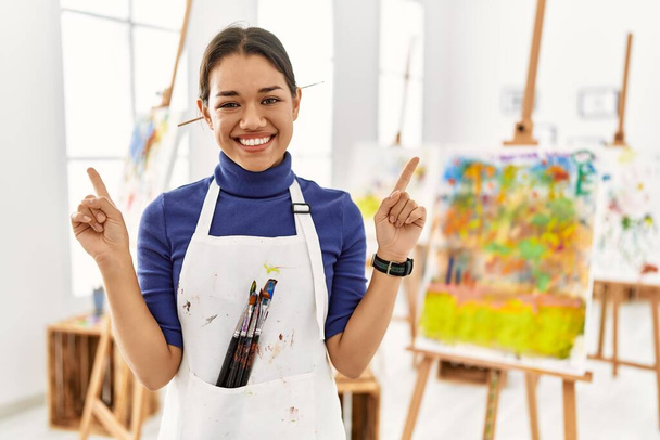 Młoda brunetka w studiu artystycznym uśmiechająca się pewnie wytykając palcami różne kierunki. miejsce na reklamę  - Zdjęcie, obraz