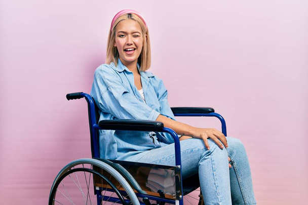 Beautiful blonde woman sitting on wheelchair winking looking at the camera with sexy expression, cheerful and happy face.  - Фото, изображение