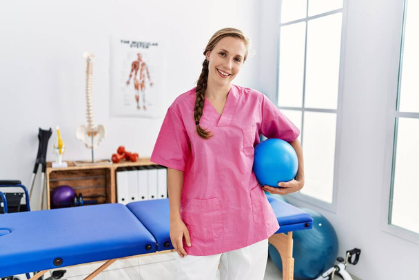Νεαρή καυκάσια γυναίκα που φοράει στολή φυσιοθεραπευτή κρατώντας μπάλα γυμναστικής στην κλινική φυσικοθεραπείας - Φωτογραφία, εικόνα
