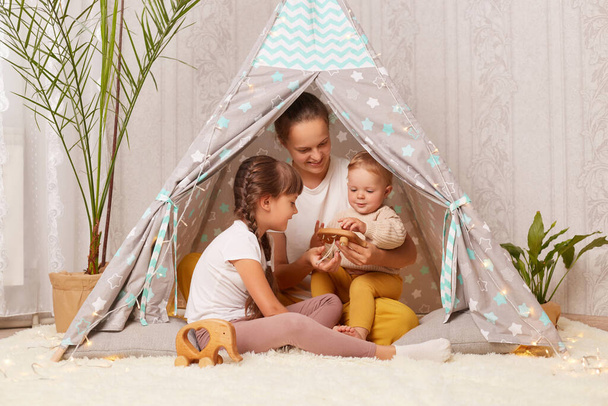 Indoor shot of mother with two kids sitting together in wigwam teepee at home, mom playing with preschool and infant children, showing wooden eco toy to her baby. - Фото, зображення