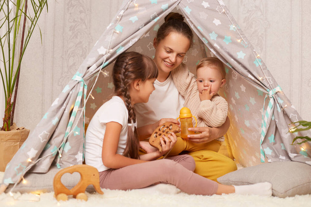 Portrait of satisfied smiling mother with two kids sitting together in wigwam, mom playing with kids in play tent, family expressing positive emotions and happiness. - 写真・画像