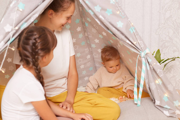 Indoor shot of family spending time at home in cozy wigwam, infant baby playing with garland lights, mother looking at her charming toddler daughter with toothy smile, elder sister sitting backwards. - 写真・画像