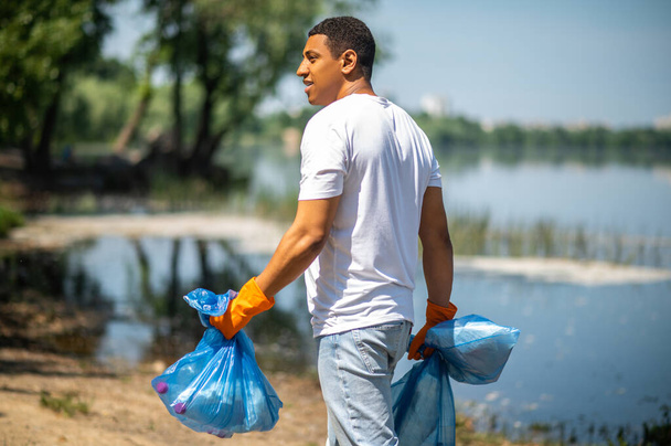 Volunteer. Side and dark-skinned view of dark-skinned smiling guy in gloves with garbage bags walking near river on fine day - Фото, изображение